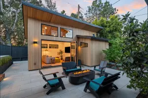 Valley Home Builders inc. Modern detached ADU with light wood exterior, glass doors, and a patio featuring a fire pit and seating amidst greenery at dusk.